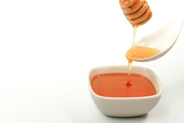 Honey is pouring from wooden honey dipper into small white bowl on white background