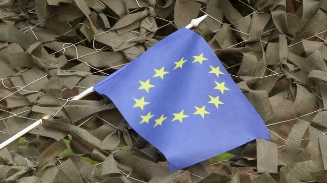 The EU flag is attached to a camouflage military net made of green fabric ribbons as support for the army