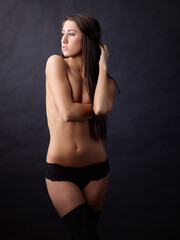 Fototapeta premium In a dark studio, a beautiful woman stands, dressed only in stockings and panties, and covers her bare breasts.
