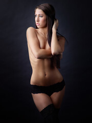Fototapeta premium In a dark studio, a beautiful woman stands, dressed only in stockings and panties, and covers her bare breasts.