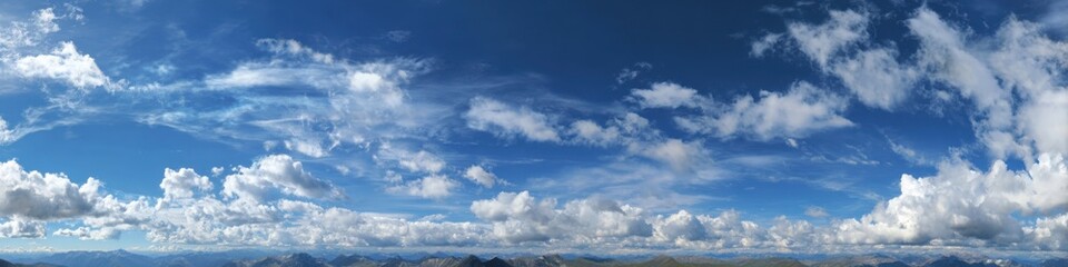 Obraz premium Panoramic view of a vast, bright blue sky dotted with fluffy white clouds. A hazy, distant mountain range is visible at the horizon