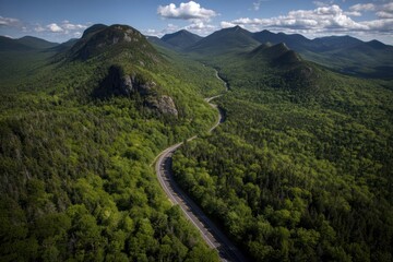 Obraz premium A winding road cuts through dense green forest and rolling mountains under a bright, partly cloudy sky