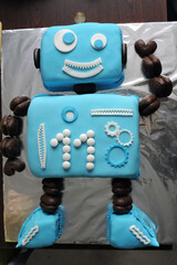 A colorful birthday cake designed in the shape of a cute blue robot. Decorated with fondant gears, buttons, wires and a lollipop at the top of its head