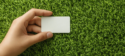 Hand holding blank white card on fresh green grass, natural background, clean and simple design, eco friendly concept, peaceful and calm atmosphere