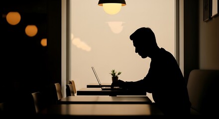 Fototapeta premium Man working on laptop in a dimly lit room with warm lighting by window