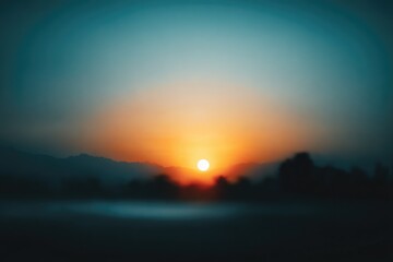 Obraz premium A serene, blurred landscape depicts a vibrant sunset over silhouetted trees and mountains, creating a peaceful and dreamy atmosphere