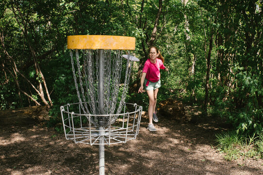 Tween girl throws putter disc at discing cage in green outdoors
