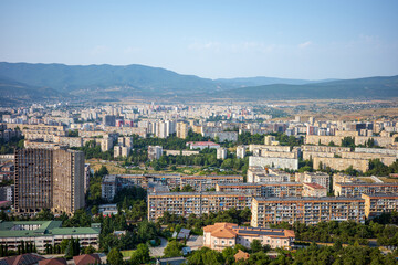 Obraz premium A wide-angle panoramic view of Tbilisi, Georgia, showcasing rows of a vibrant Soviet-era apartment blocks, modern high-rise buildings, and lush greenery framed by the Caucasus mountains