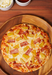 Tasty pineapple and ham hawaiian pizza flatbread baked on wooden plate, delicious melting mozzarella cheese on wooden board, homemade cooking ingredients lying on dark background, food advertisement.