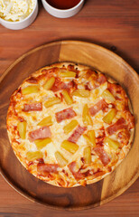 Tasty pineapple and ham hawaiian pizza flatbread baked on wooden plate, delicious melting mozzarella cheese on wooden board, homemade cooking ingredients lying on dark background, food advertisement.