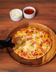 Tasty pineapple and ham hawaiian pizza on wooden plate using pizza cutter to slice delicious melting mozzarella cheese on board homemade cooking ingredient lying on dark background, food advertisement