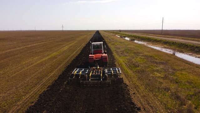 An aerial view of a red tractor plowing a long, straight furrow of dark earth