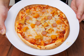 Tasty pineapple and ham hawaiian pizza baked on baking steel plate, delicious melting mozzarella cheese on wooden board, homemade cooking ingredients lying on dark background, food advertisement.
