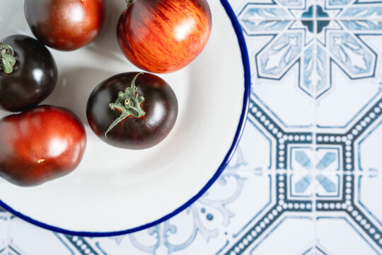 Ripe red and black beauty tomatoes on white plate with blue rim. Tiled kitchen table. Cooking with copy space