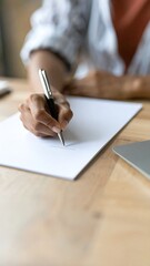 Person writing on paper at a wooden desk with laptop