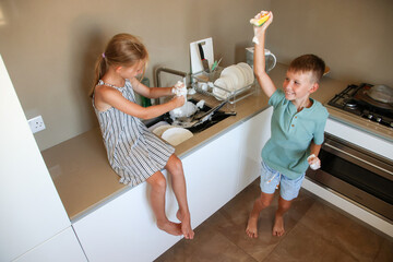 Happy children playing with soap foam and sponge while washing dishes in kitchen