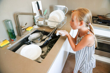 Little girl blowing soap foam bubbles while helping wash dishes in modern home kitchen
