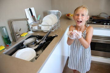 Happy little girl smiling and playing with soap foam while helping wash dishes at sink