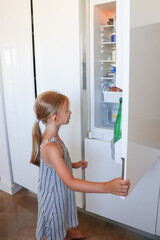 Little girl opening refrigerator door and looking inside modern white kitchen at home