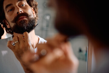 Reflection in mirror of man styling his beard with comb.