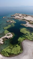 Aerial view of a rocky coastline with a shallow lagoon, turquoise water, and light green vegetation under a clear blue sky