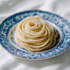 A plate of creamy, pale yellow spaghetti, neatly coiled and topped with grated cheese, sits on a white fabric background. The plate features a blue and white floral pattern