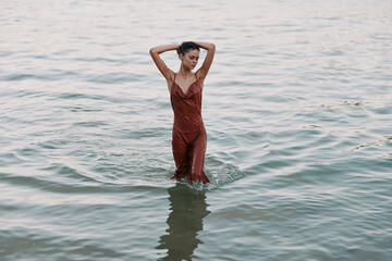 A woman stands waist deep in calm water, wearing a rust red sleeveless jumpsuit, hands raised behind her head, creating a serene, summery moment by the sea.
