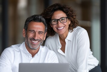 Smiling professional couple in a bright office, the woman wears glasses and leans on a man, both in white shirts, happy and at ease