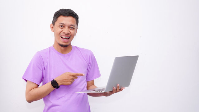Happy asian man wearing purple t-shirt and holding laptop computer working online and looking at camera isolated on white background, online education
