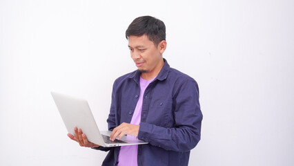Happy asian man wearing purple t-shirt and holding laptop computer working online and looking at camera isolated on white background, online education