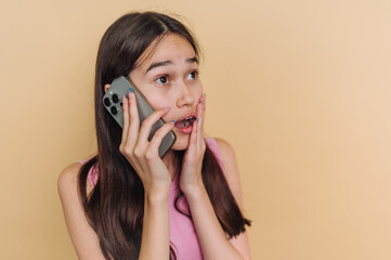 Fototapeta premium portrait of young girl showing a shocked expression holds her smartphone to her ear on beige background