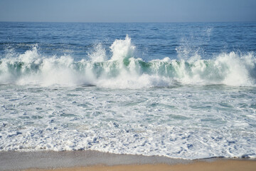 A closeup shot of the ocean with wind waves crashing onto the shore, creating a beautiful pattern of furrows in the water. The landscape is enhanced by cumulus clouds in the sky