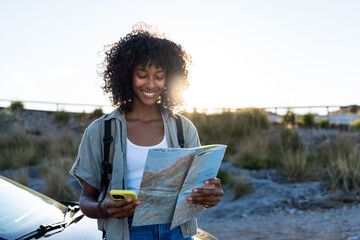 Smiling woman with a backpack standing next to her car, holding a map and smartphone, planning her route during sunrise in the Balearic Islands. Concept of hiking, travel and exploration. © Martí Rosselló