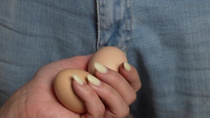 Two raw chicken eggs in a woman hand between a man legs in blue jeans, close up. Eggs in hand as a symbol of male testicles and problems with them