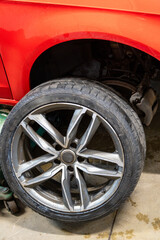 Winter to Summer Tire Change Service with Removed Wheel Leaning on Red Vehicle in Garage Workshop. Seasonal Tire Replacement Process Showing Winter Tread Pattern on Dismounted Tire with Hydraulic Jack