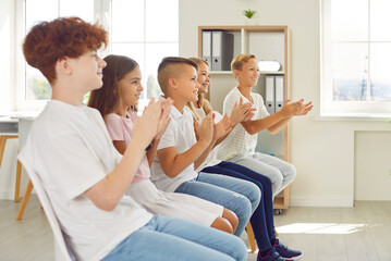 Happy children sitting in a row on chairs and clapping hands at a school event in classroom. Group...
