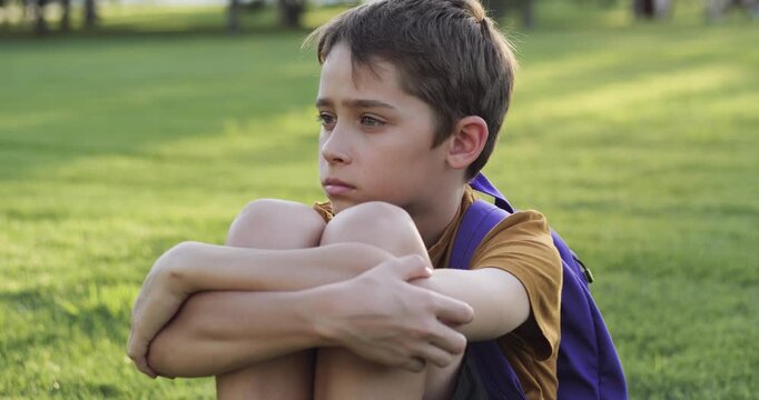 Child psychology concept: portrait of child sitting on green grass in park, hugging knees. Thoughtful boy with calm, pensive face looks ahead. mental health, mindfulness, and emotional children - Powered by Adobe