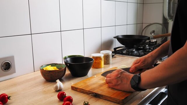 In a modern kitchen, a person prepares to cook, chopping garlic and cherry tomatoes while surrounded by herbs, spices, and cheeses. A skillet waits on the stove for a delicious dish