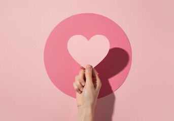 A hand holding a paper heart in front of a pink circle, casting a shadow.