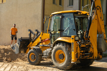 Two construction workers at a sunny building site: one operates a yellow backhoe loader, the other assists with a shovel for earthmoving.