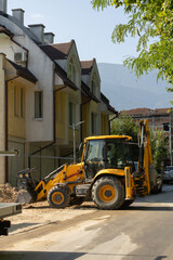 Bright yellow backhoe loader on dirt path by asphalt road. Modern residential buildings in background suggest urban/suburban construction/development.