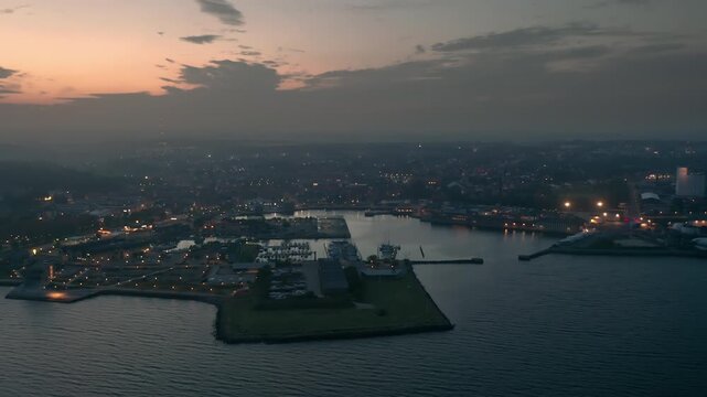 Aerial night view of Thisted, Denmark. Town lights, marina, Limfjord coastline, and surrounding neighborhoods create a tranquil Nordic cityscape under a twilight sky