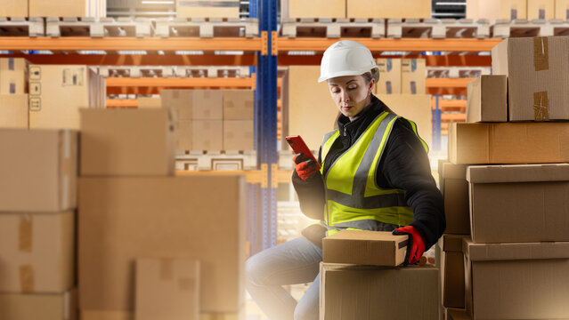 Logistics and warehouse work, female worker in safety vest and helmet scanning packages with mobile device among stacked cardboard boxes, shipping, delivery, storage.