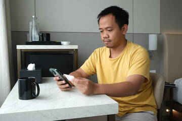 A man in a yellow shirt shops online using his smartphone and credit card, sitting at a marble desk in a modern room.  
