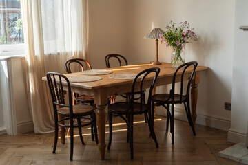 Spacious dining room interior with wooden farmhouse table and bentwood chairs in vintage style, vintage table lamp and vase with summer flowers. Aesthetic cozy interior with warm light