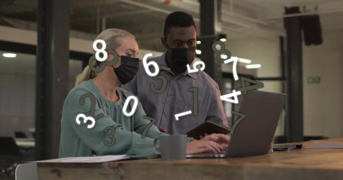 Working masked coworkers analyzing data in open office with laptop, tablet, coffee mug papers