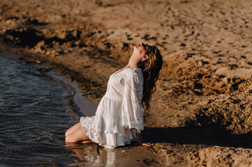 Photograph, portrait of a beautiful curly woman sitting on the sand on the beach against the backdrop of the sea, ocean in a white sundress, dress at sunset.