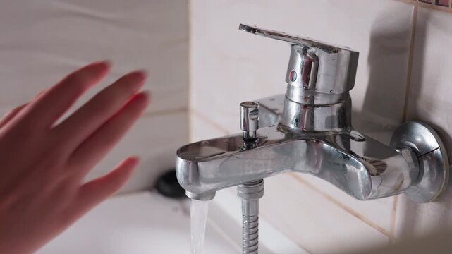 Hand view lady turning on shower tap, on chrome bath mixer, fingers twist knob, hose visible, soft light across tiles, bathroom setting, routine hygiene start, calm motion before washing