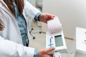 Female doctor examining electrocardiogram paper result in hospital, performing medical activity using portable medical device for electrocardiogram test