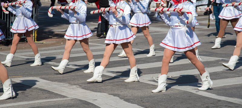 Panorama view parade performers in white skirts, red scarves march oversized J shaped candy cane props in Dallas. Crosswalk setting, blurred crowd evoke urban festivity, choreographed tradition